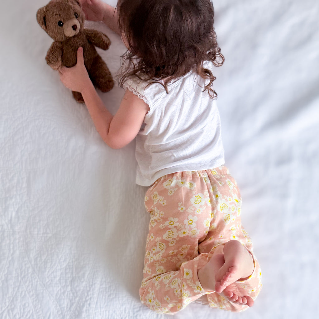 A toddler lies on a white textured blanket, holding a brown teddy bear. The child is wearing a white short-sleeve top and peach pants with a small yellow and white floral pattern.
