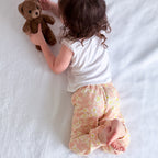 A toddler lies on a white textured blanket, holding a brown teddy bear. The child is wearing a white short-sleeve top and peach pants with a small yellow and white floral pattern.