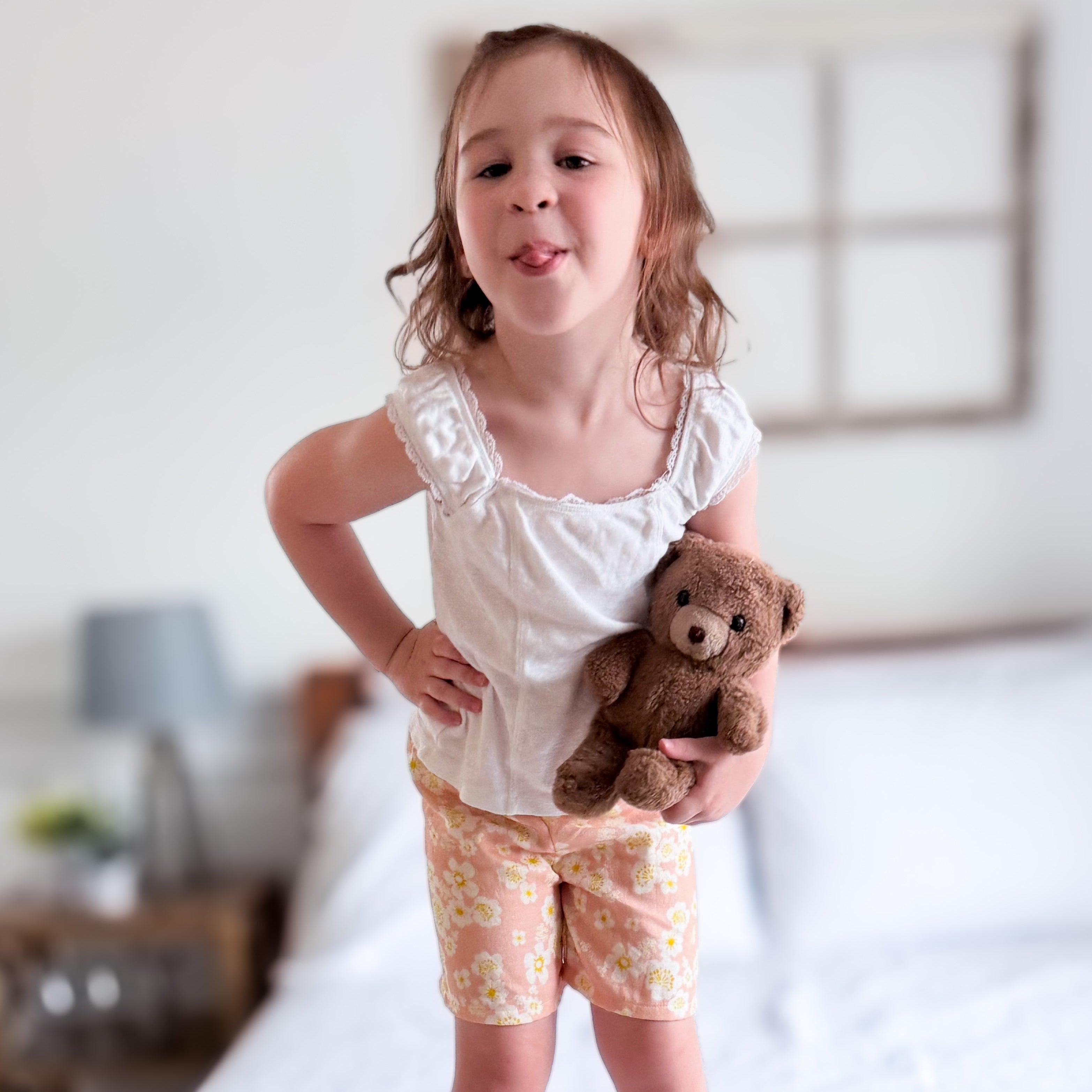 A young girl with light brown hair stands barefoot on a white bed, wearing a white ruffled short-sleeve top and peach floral shorts, while holding a brown teddy bear.